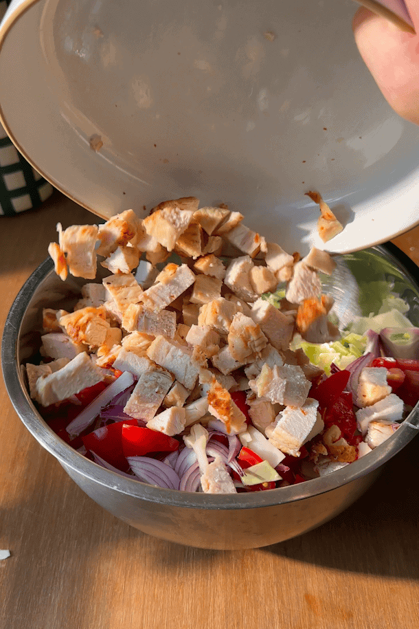 Diced cooked chicken is being poured from a bowl into a metal bowl filled with sliced onions, tomatoes, and lettuce, all on a wooden surface in bright sunlight.