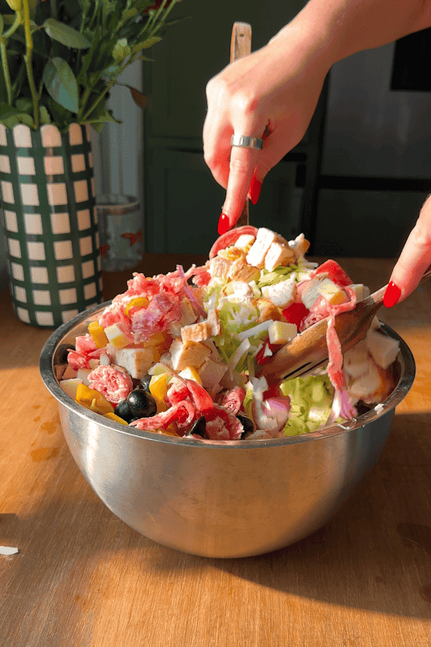 A person with red nails mixes a colorful chopped salad in a metal bowl on a wooden table. A vase of flowers is in the background.