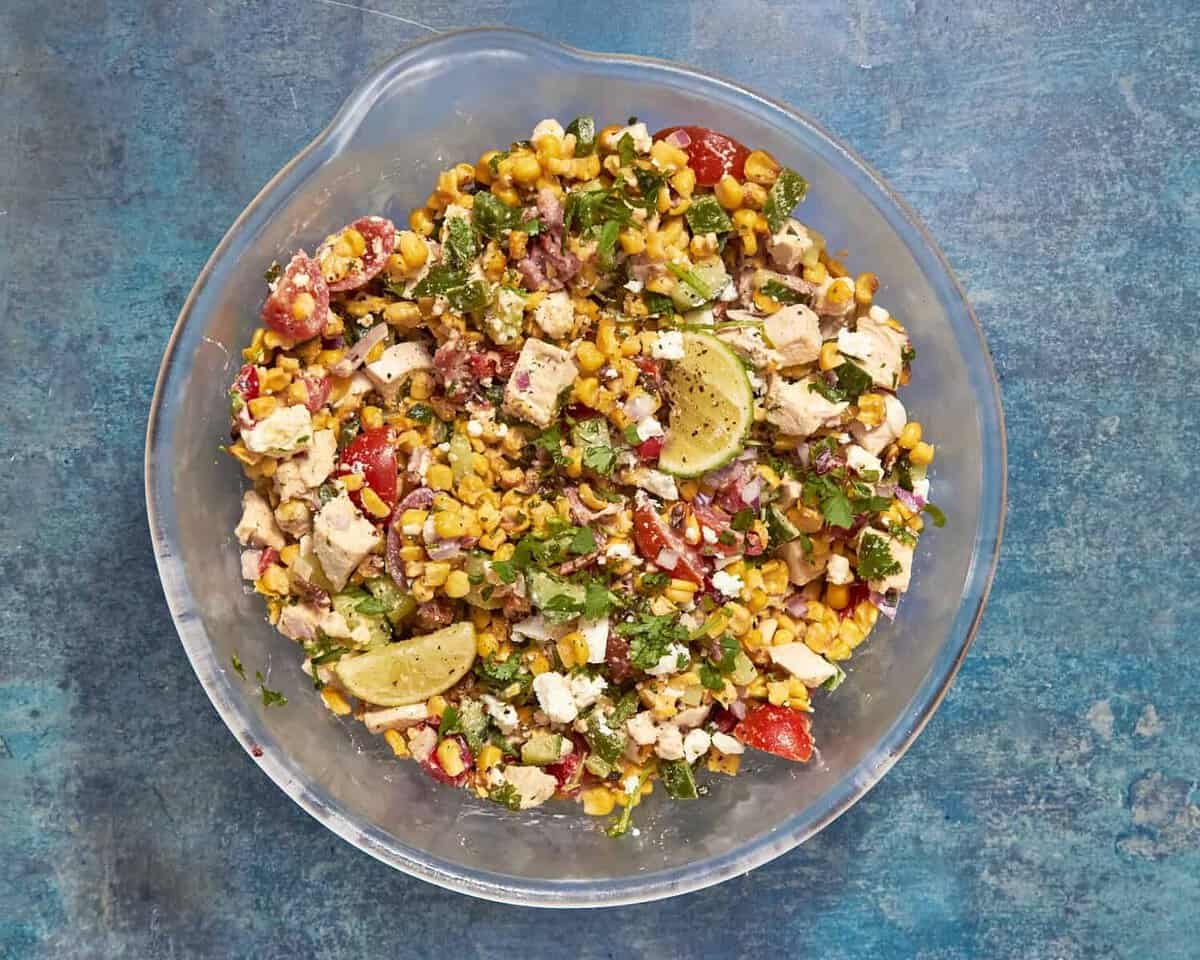 A glass bowl filled with a colorful salad containing roasted corn, chopped chicken, tomatoes, red onions, cilantro, feta cheese, and lime wedges, set on a blue textured surface.