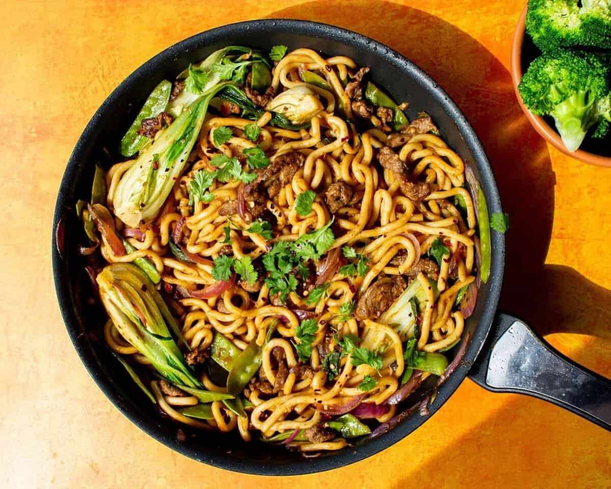 Beef udon noodles in a large pan with pok choi and garnished with coriander.