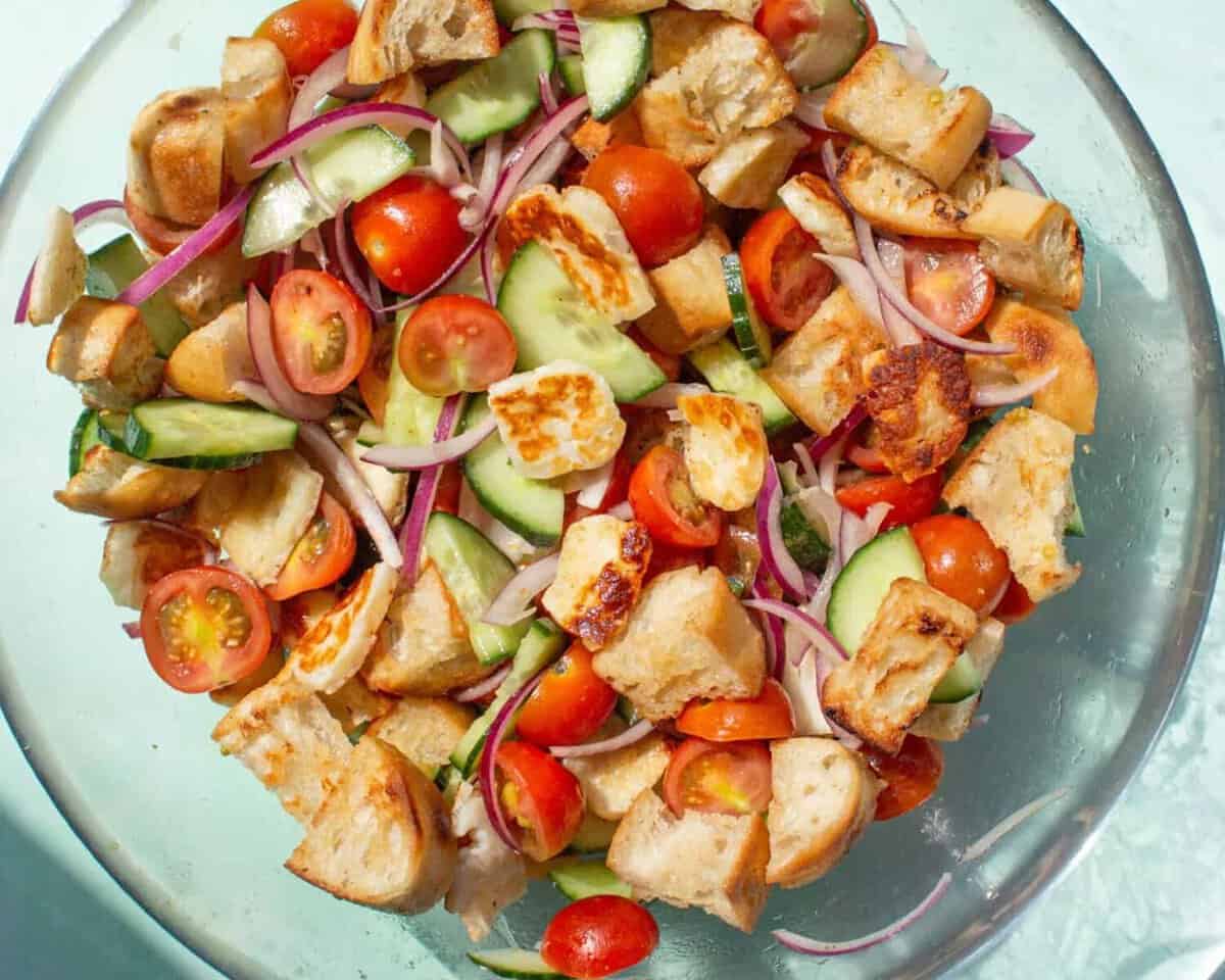 Salad with cherry tomatoes, red onion, bread pieces and fried halloumi in a glass bowl.