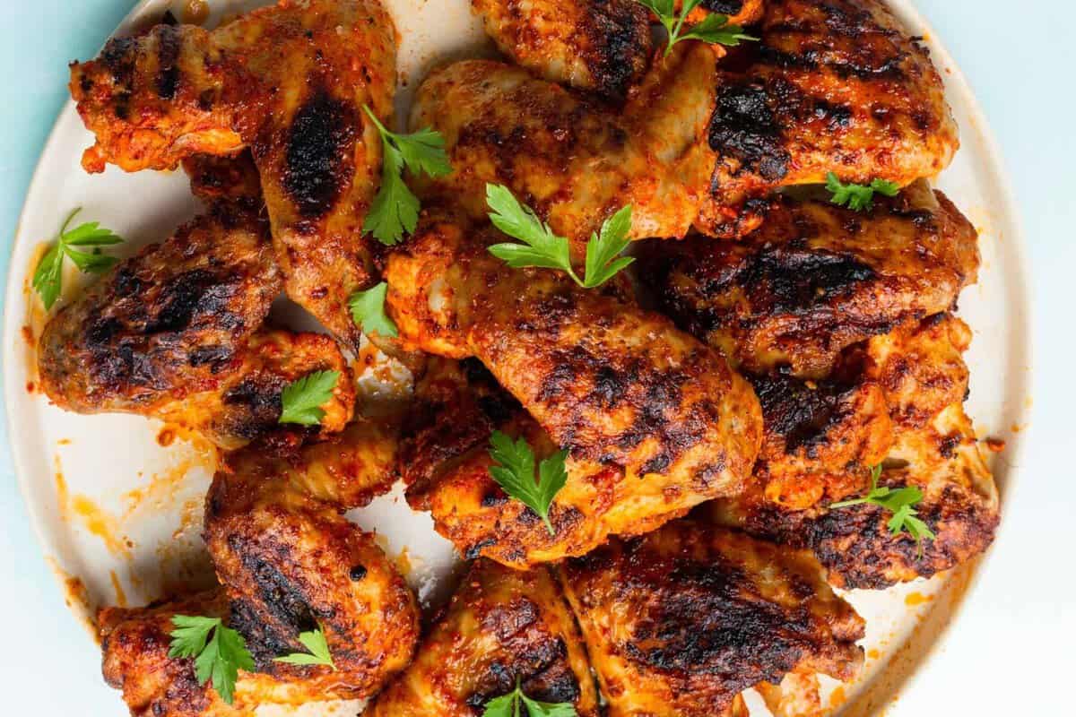A plate of grilled chicken wings with charred, crispy skin, garnished with fresh parsley leaves. The wings are coated in a reddish seasoning.