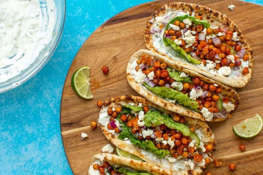 Three flatbread wraps filled with lettuce, roasted chickpeas, chopped red onion, and crumbled cheese sit on a wooden board with lime wedges, next to a bowl of white sauce, on a bright blue background.