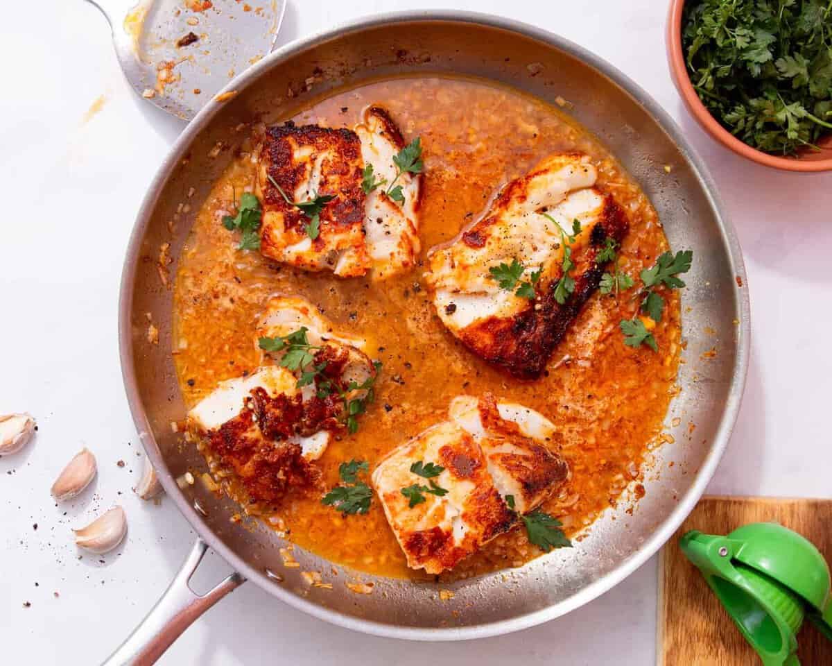 4 pieces of browned white fish in a large pan in an orange coloured sauce with some fresh parsley.