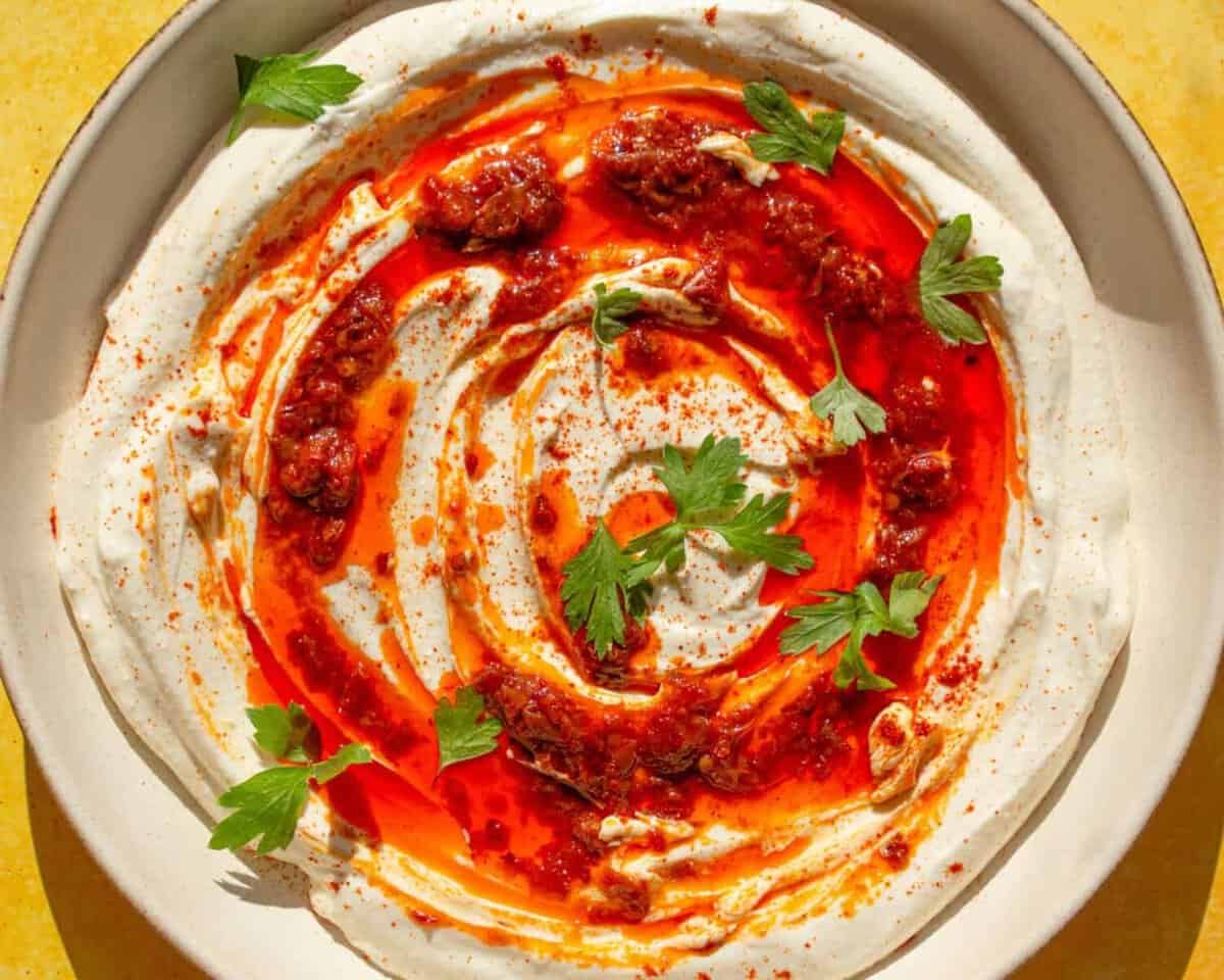 A bowl of creamy yogurt dip is swirled with bright red chili oil and garnished with fresh parsley leaves. The dip sits on a white plate, and the colors are vibrant and appetizing.
