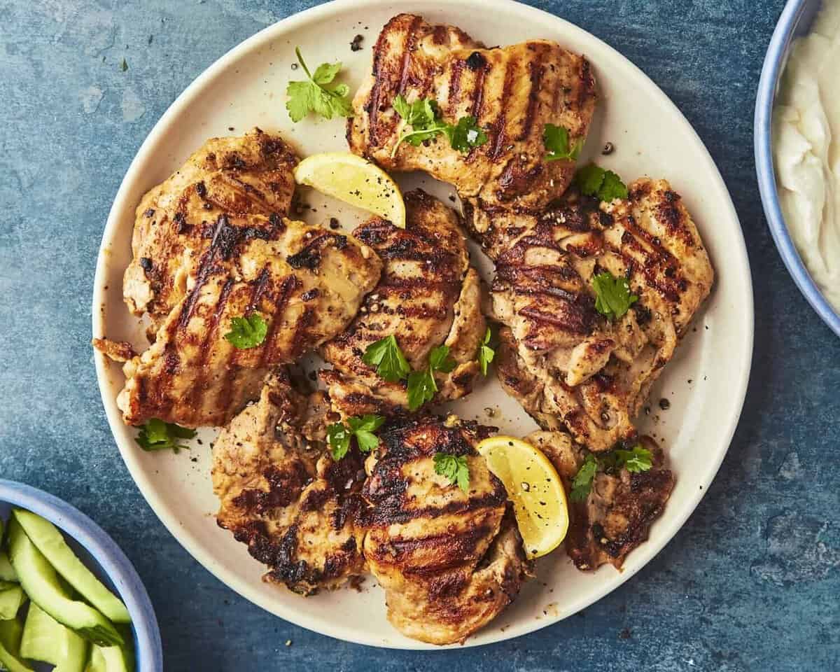 Grilled chicken thighs with char marks, roasted corn, and fresh cilantro, garnished with lemon wedges on a round beige plate set on a blue surface. Sliced cucumbers and a dip are partially visible in nearby bowls.