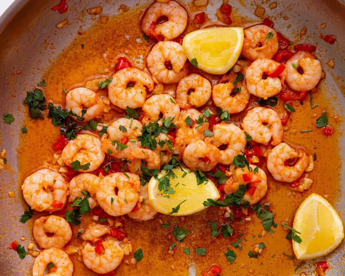 A large pan with King prawns in a sauce with lemon wedges, fresh parsley and red chillies.