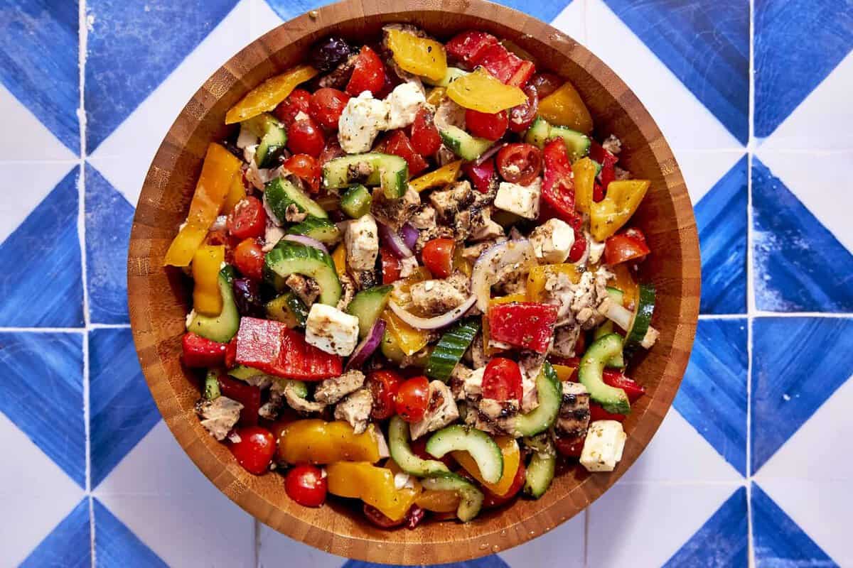 A wooden bowl filled with colorful Greek chicken salad, including tomatoes, cucumbers, red onions, bell peppers, olives, and feta cheese cubes, sits on a blue and white tiled surface.
