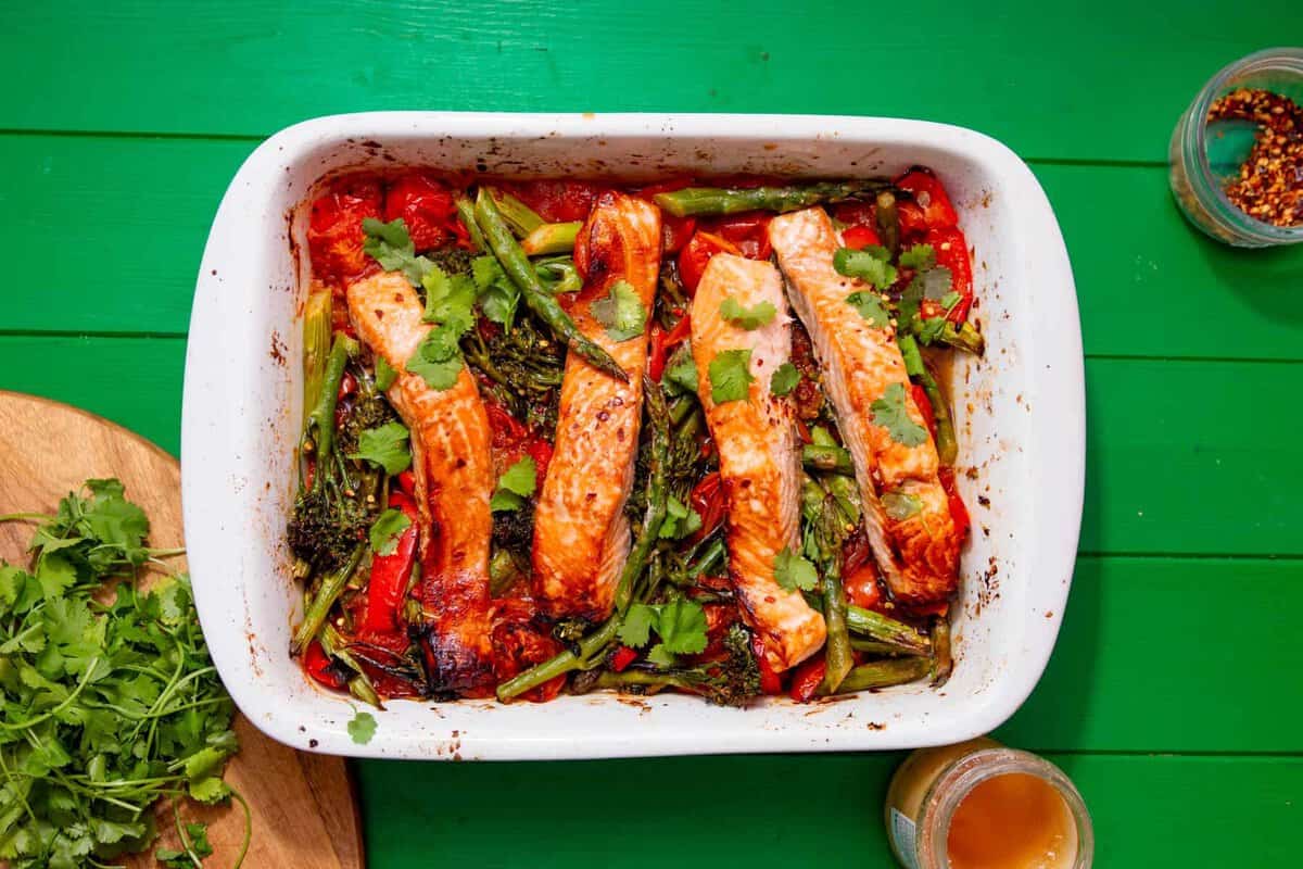A white baking dish on a green table holds roasted salmon fillets with asparagus, tomatoes, and fresh cilantro. Fresh cilantro and small jars with seasonings are nearby.