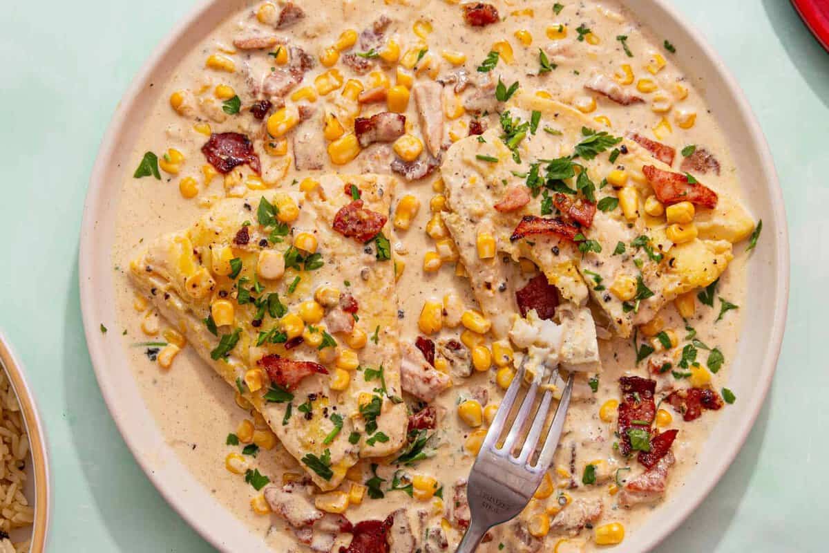 A plate of fish fillets topped with creamy sauce, corn, chopped bacon, and parsley, with a fork taking a bite. The dish is colorful and garnished with herbs.