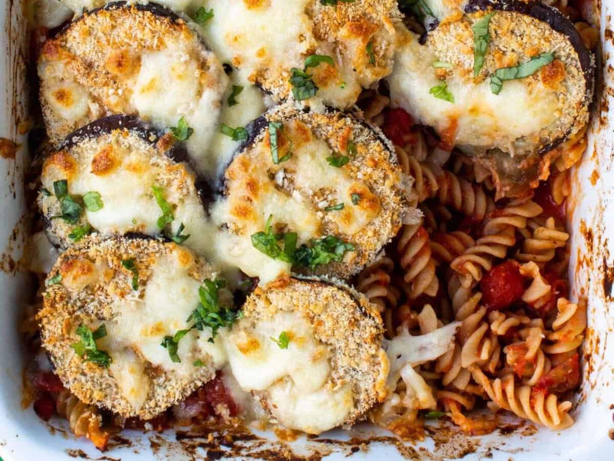Aubergine parmigiana pasta bake with aubergine round discs, golden brown mozzarella/parmesan and a portion taken. showing the tomatoey pasta.
