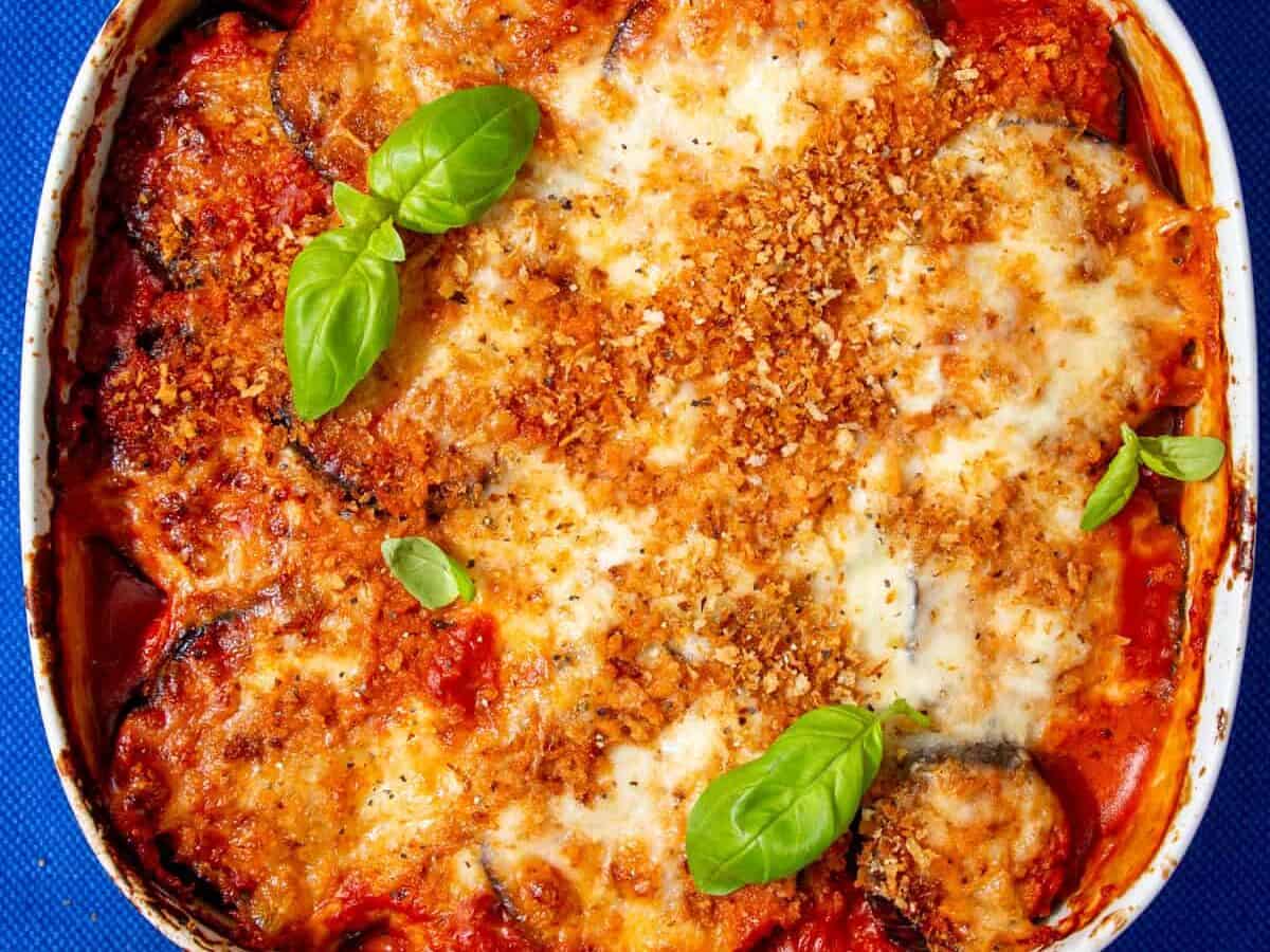 A white square dish with aubergine parmigiana topped with a golden browned cheesy topping and browned breadcrumbs next to a wooden board with grated cheese and basil leaves next to a little bowl of peppercorns.