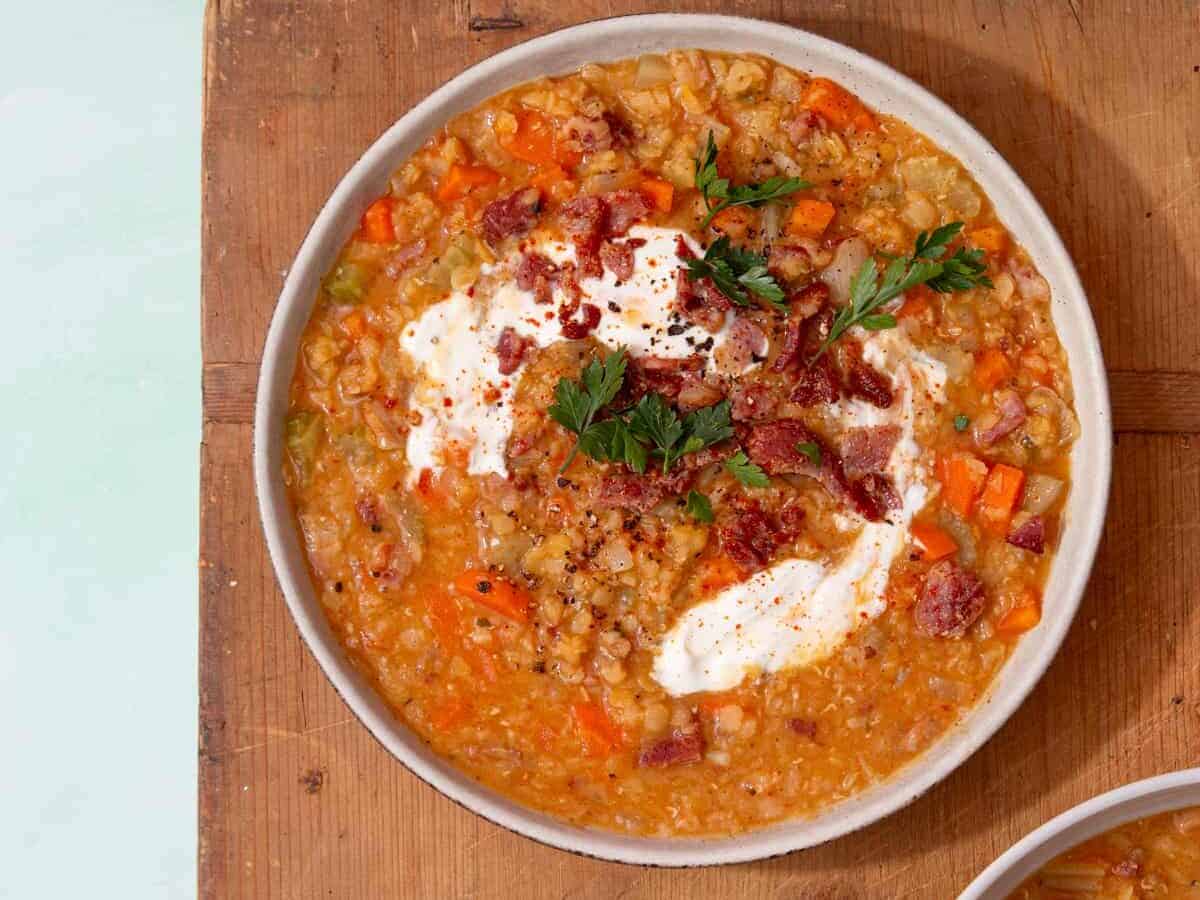 A bowl filled with lentil soup topped with bacon and sour cream on a wooden board with another 2 bowls in partial view.