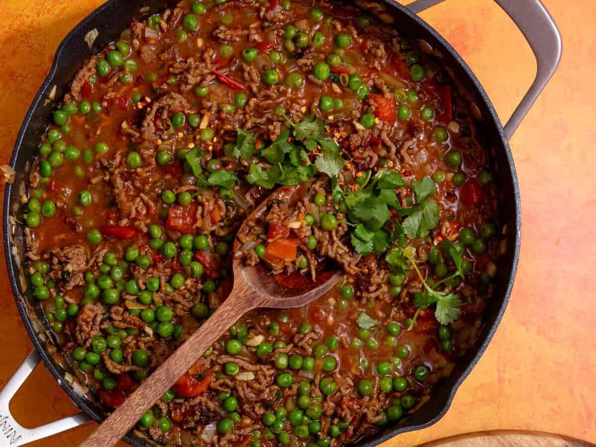 A large pan with mince curry with peas topped with coriander with a wooden spoon with naan breads.