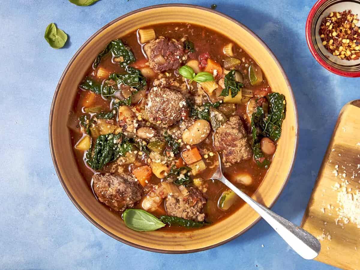 A bowl of hearty Beef Minestrone Soup with meatballs, beans, leafy greens, and chopped vegetables on a blue surface. Fresh basil leaves and a pot of soup are nearby, along with a small bowl of red pepper flakes.