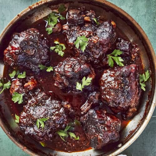 Overhead shot of braised oxtails in a rich glossy sauce, scattered with fresh parsley.