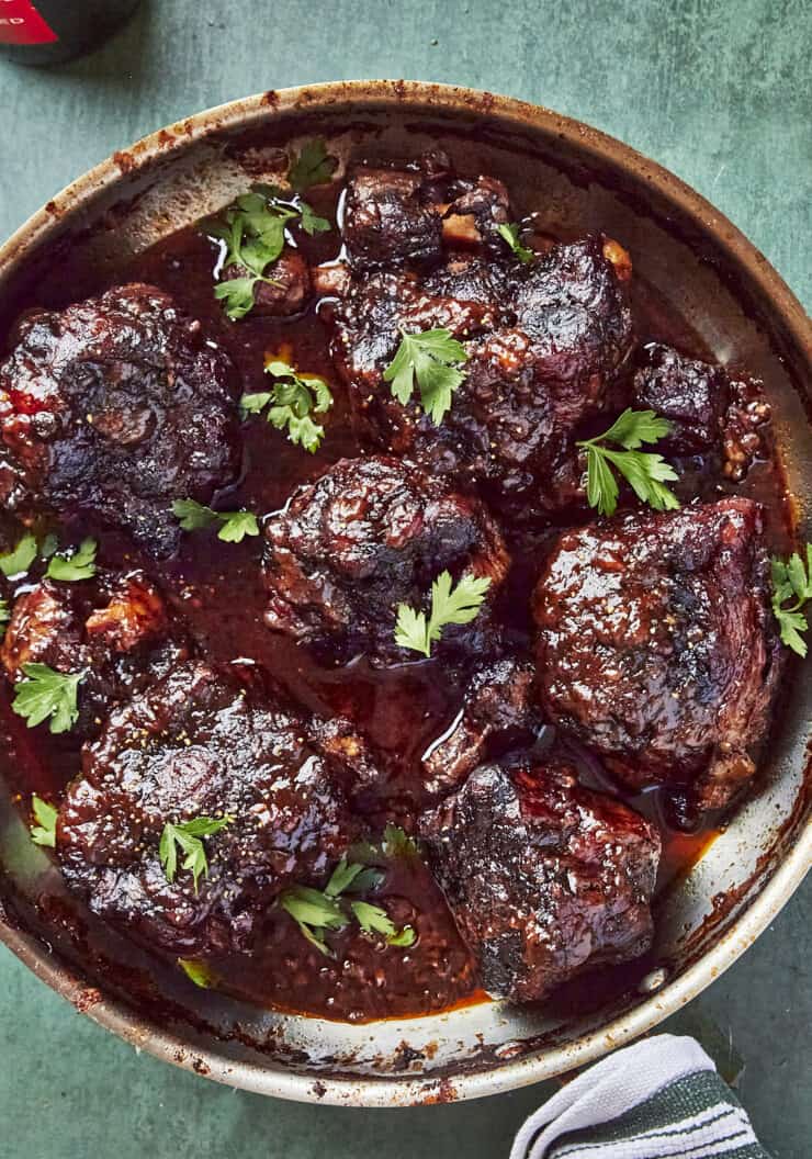 Overhead shot of braised oxtails in a rich glossy sauce, scattered with fresh parsley.