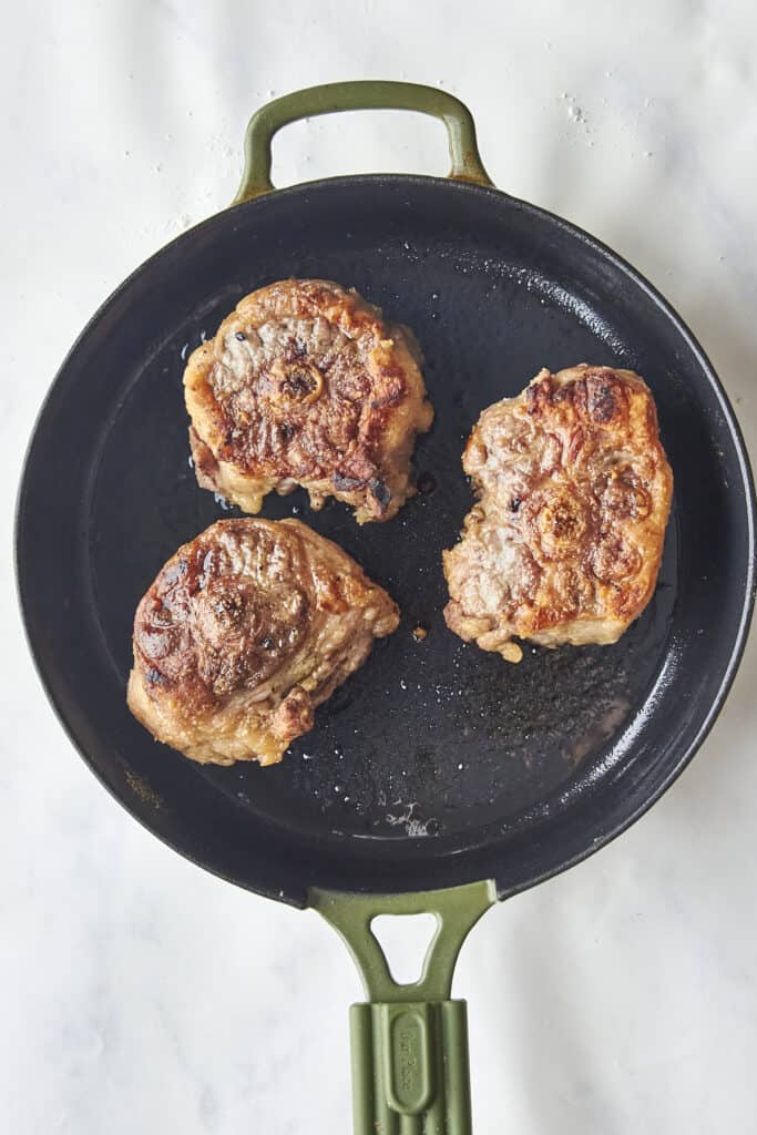 Three browned steaks are cooking in a black frying pan with a green handle, resting on a white countertop.