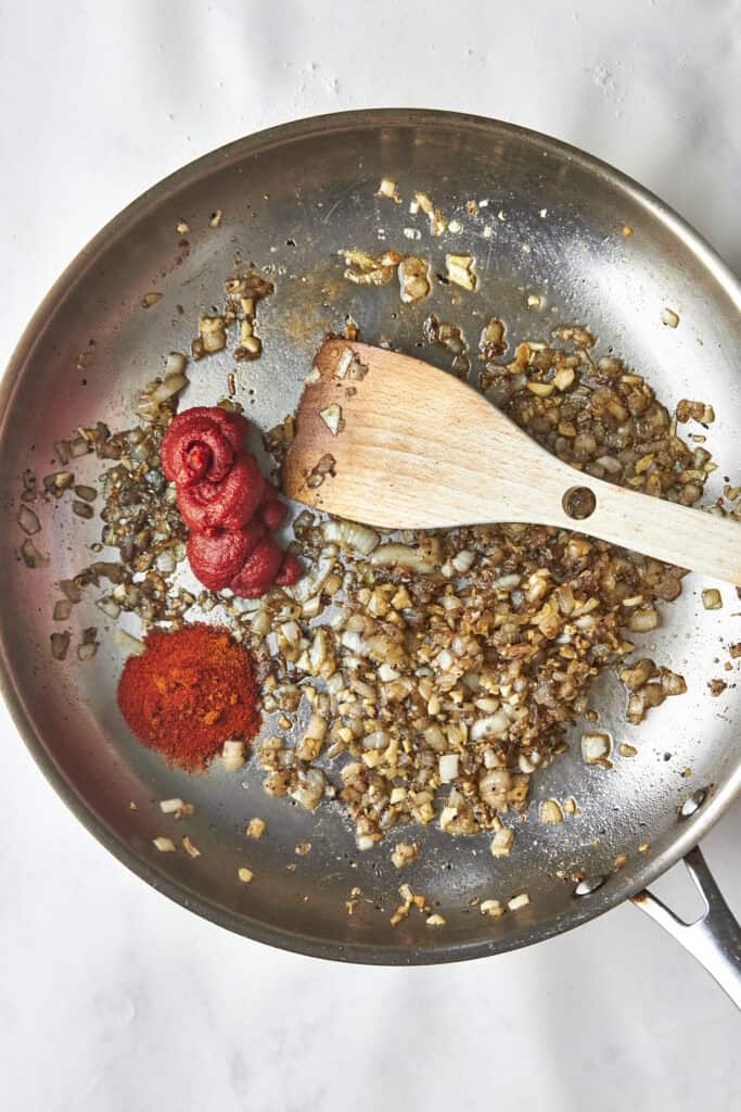 Chopped onions sautéing in a pan with a wooden spatula, next to piles of paprika and tomato paste on a white countertop.