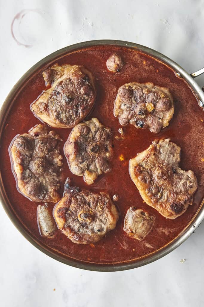 A pan filled with pieces of browned meat simmering in a rich red sauce, viewed from above on a light surface.