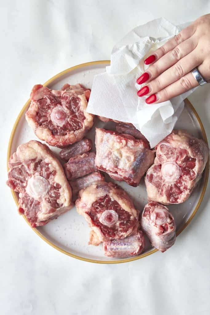 A hand with red nails pats raw oxtail pieces dry with a paper towel on a white plate.