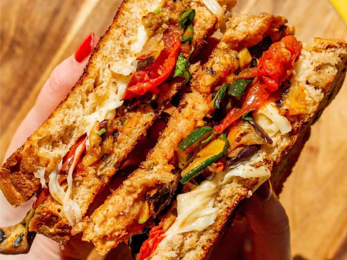 Side view of cheesy toastie held with 2 hands showing filling of roasted courgette slices, tomatoes and melted mozzarella.