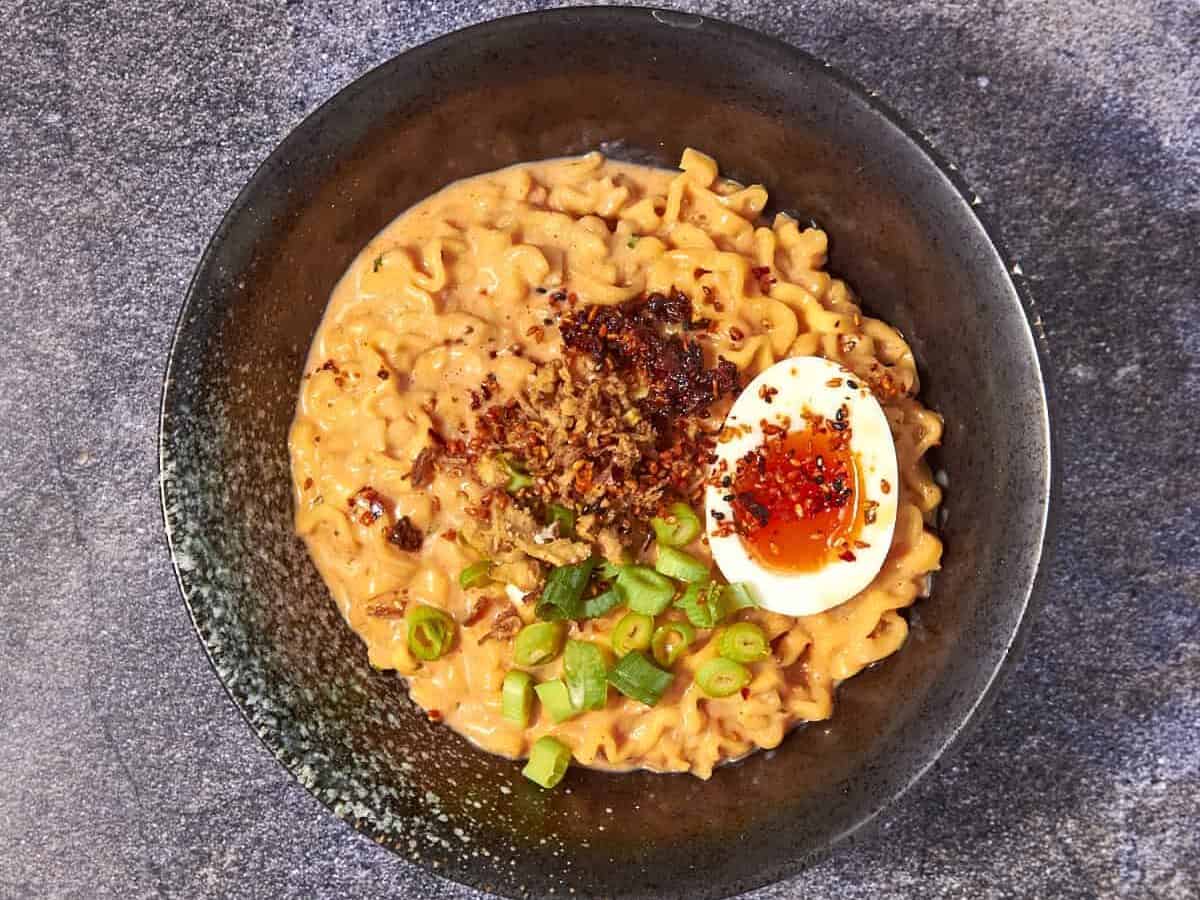 A bowl of creamy buldak noodles topped with a halved soft-boiled egg, chopped green onions, chili flakes, and fried onions, with wooden chopsticks placed above the bowl on a stone surface.