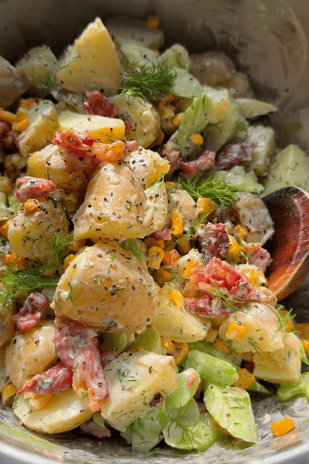 Creamy dill potato salad mixed together in a bowl with a wooden spoon, showing potatoes, bacon, sweetcorn, celery and dill dressing combined.