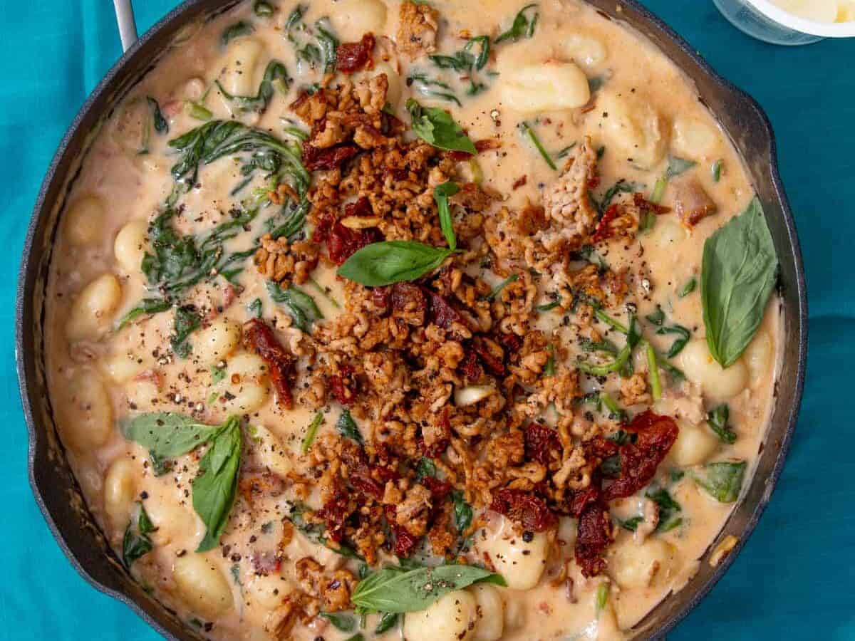 Gnocchi with a creamy sauce with pork mince, spinach and sun-dried tomatoes topped with basil in a pan in a blue background.