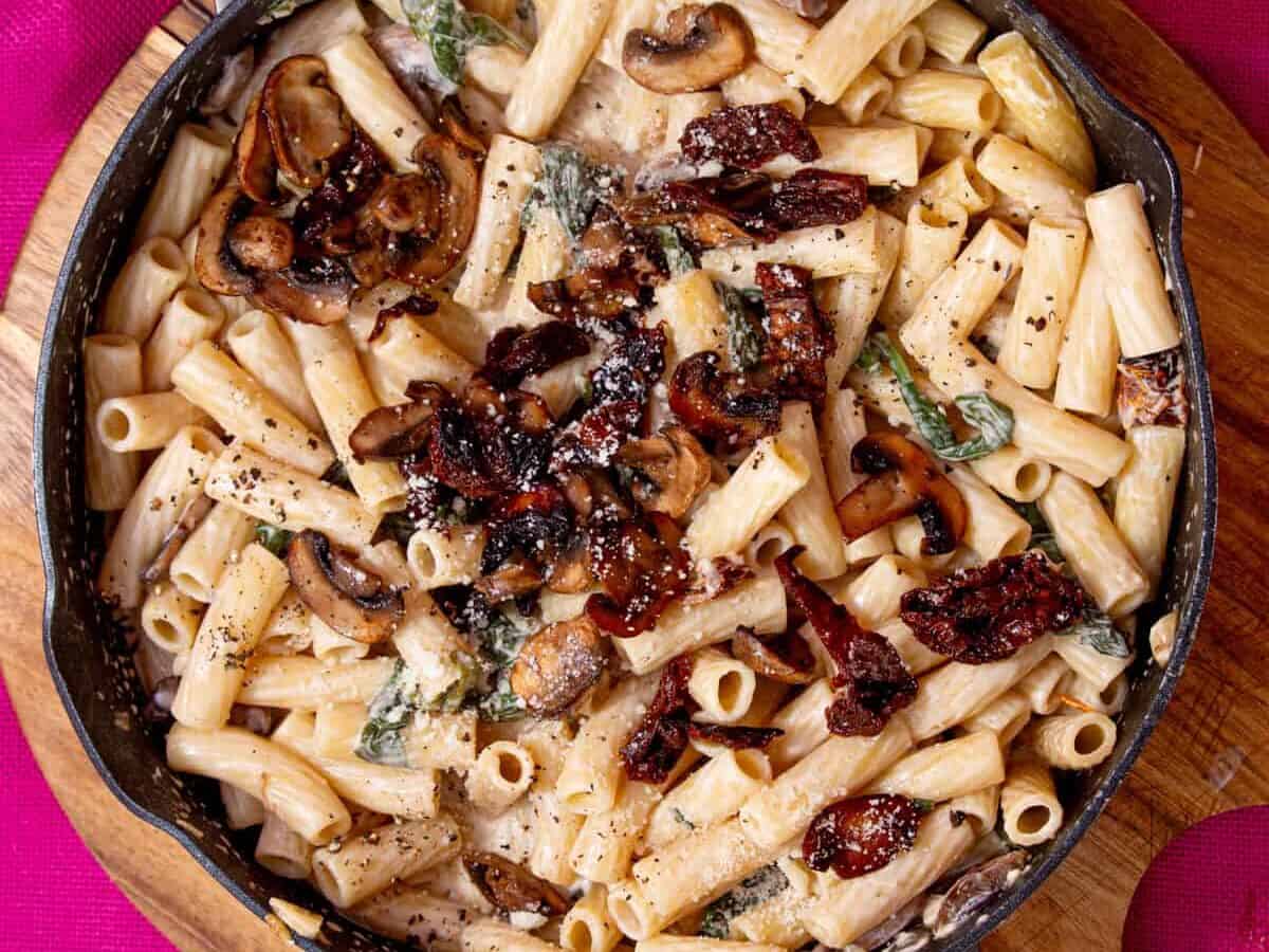 A large pan with a creamy mushroom pasta topped with mushrooms and sun-dried tomatoes on a pink back ground.