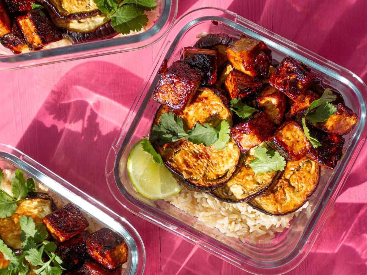 Crispy soy tofu, aubergine & rice in 4 meal prep containers topped with coriander and a wedge of lime on a pink background.