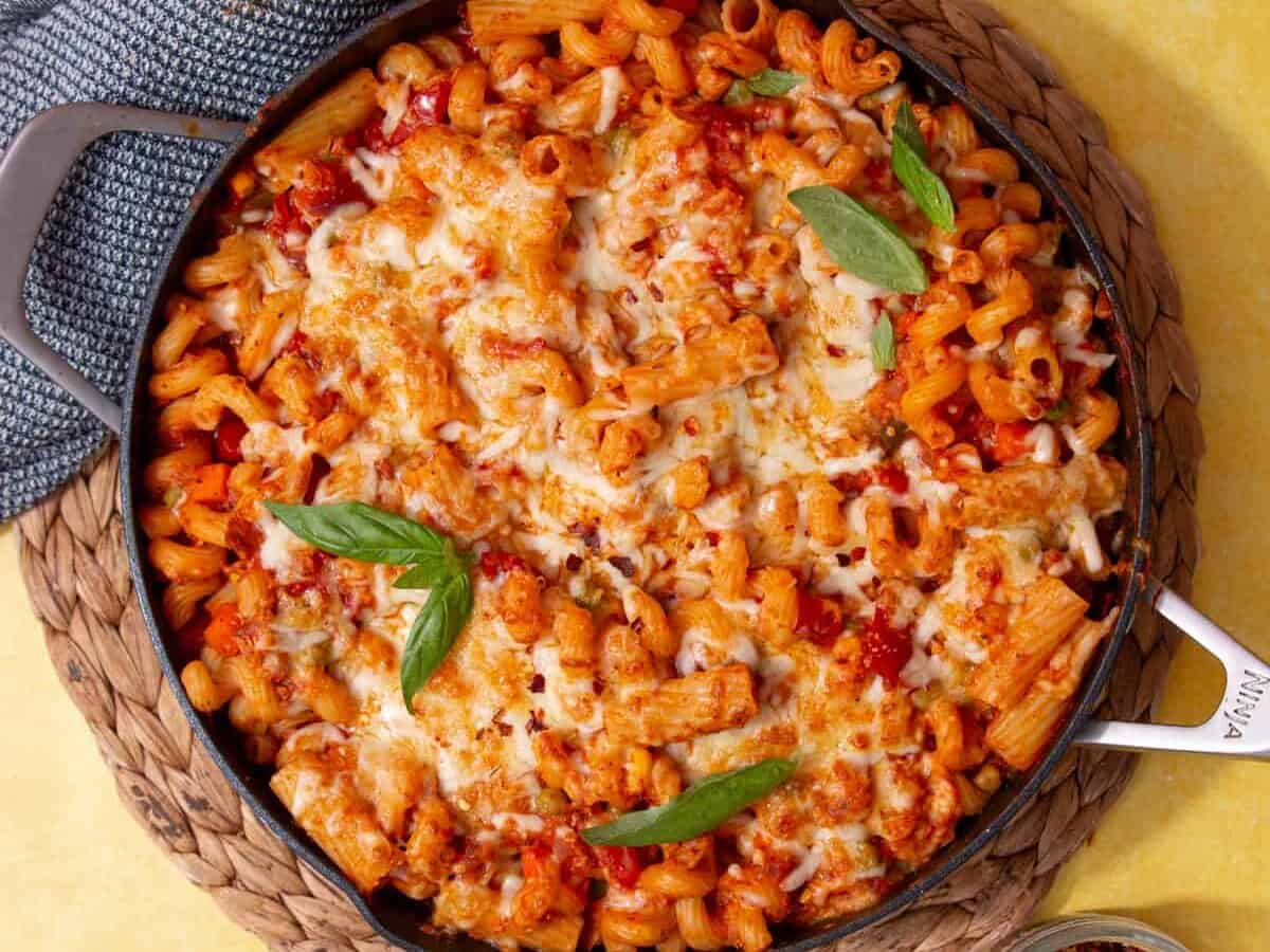 Baked pasta in a skillet topped with melted cheese, tomato sauce, and fresh basil leaves, set on a woven placemat with a gray towel nearby.