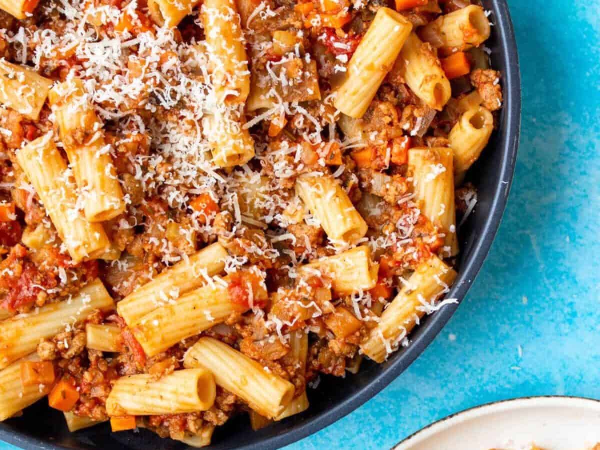 A pan with bolognese with rigatoni pasta and parmesan shavings with a bowl of bolognese pasta.