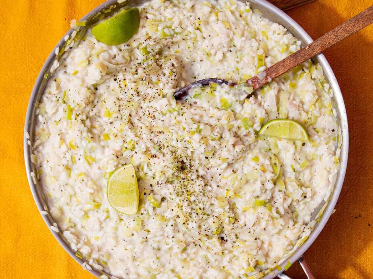 A large stainless teel pan with fish risotto topped with lime wedges, stirred with a wooden spoon on a mustrad background with a plate with leeks in partial view.