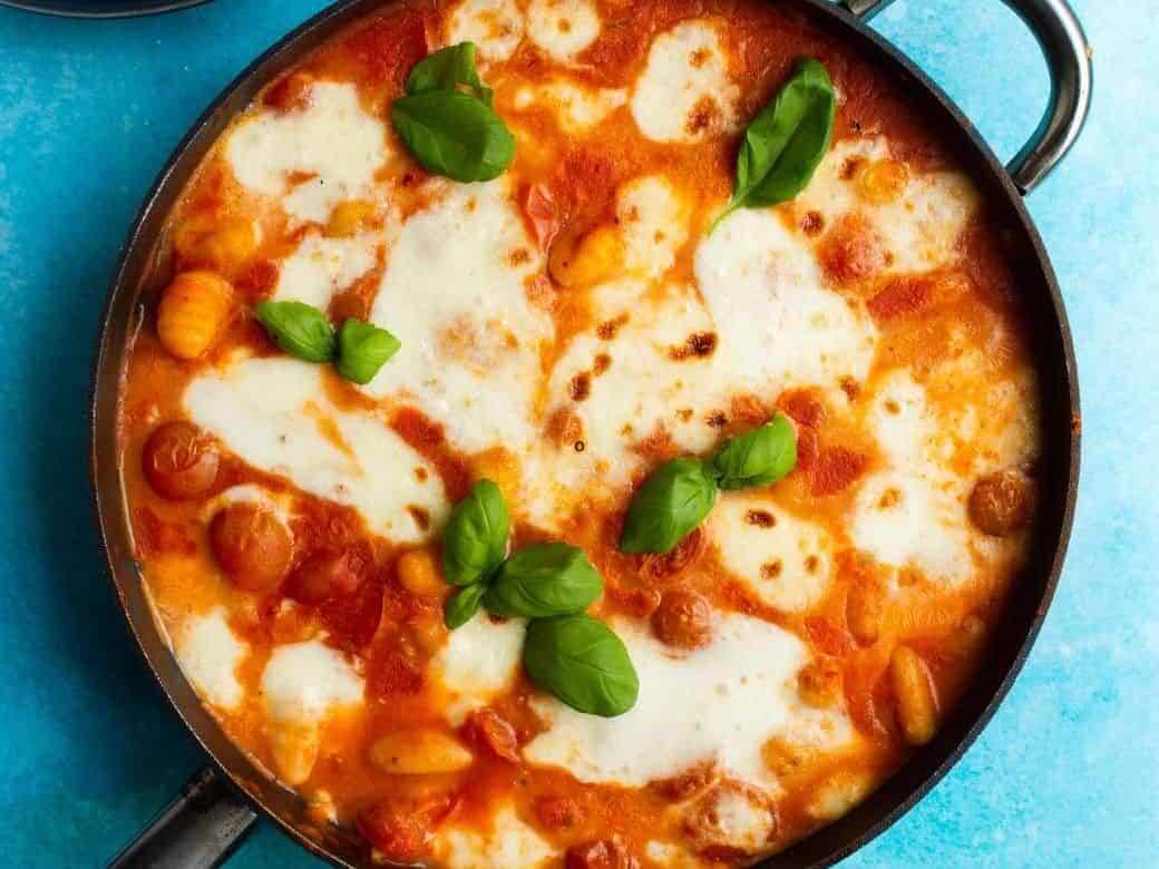 Gnocchi Alla Sorrentina in a pan topped with fresh basil on a blue background with bowls of veg.