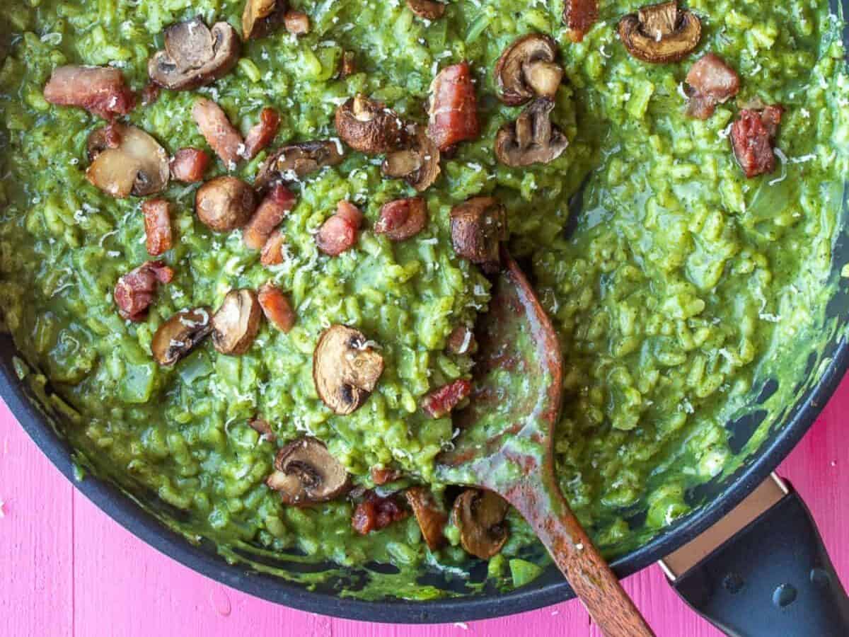A large pan with green risotto topped with bacon pieces, mushrooms and parmesan cheese.