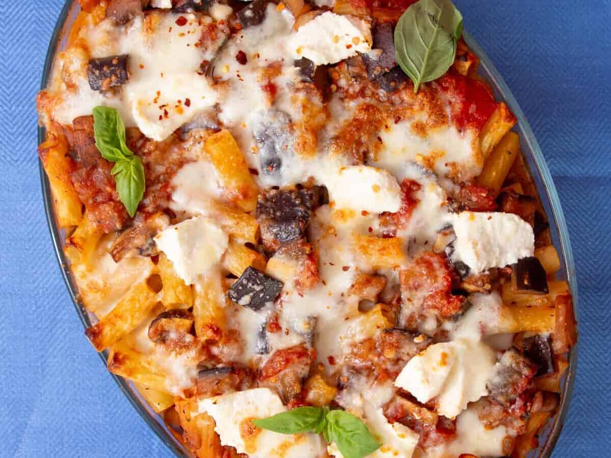 An oval dish filled with baked rigatoni pasta and a cheesy browned topping garnished with fresh basil on a blue background.