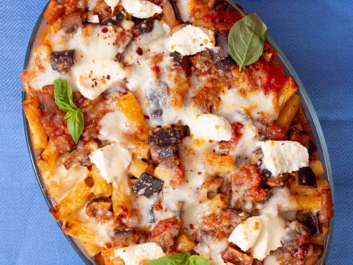 An oval dish filled with baked rigatoni pasta and a cheesy browned topping garnished with fresh basil on a blue background.