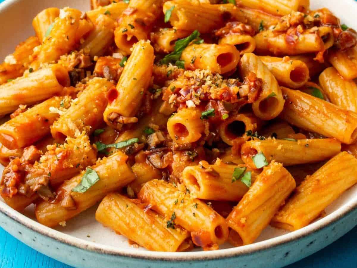 A bowl of Mushroom Rigatoni pasta with breadcrumb topping and fresh basil with a small bowl of breadcrumbs and the pan in partial view.