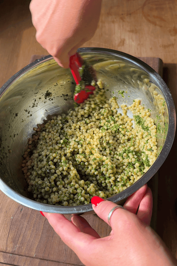 Cooked giant couscous being tossed with salsa verde dressing in a large bowl.