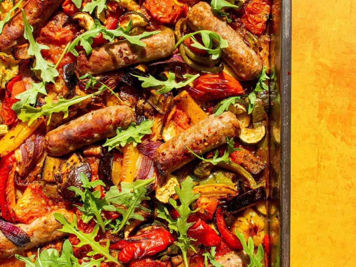 Roasted vegetables and sausages in a tray garnished with rocket on a yellow background.