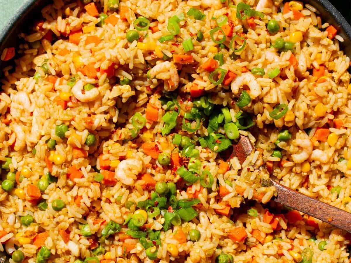 A pan with cooked rice and vegetables and prawns topped with spring onions and chops stick in pan and a box of them next to pan.