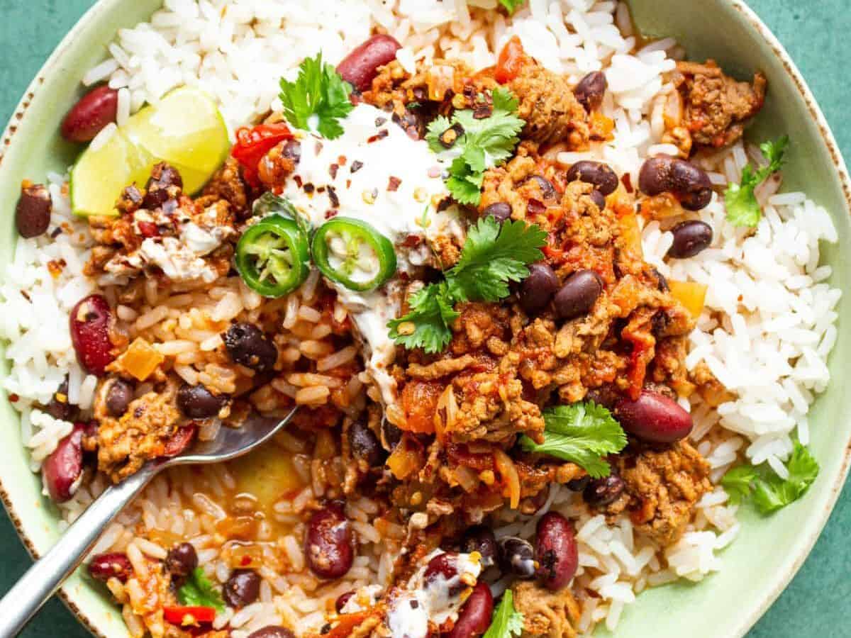 Slow cooker chilli con carne on a bowl on a bed of rice garnished with coriander and green chillies, partial view of chilli flakes and rice.