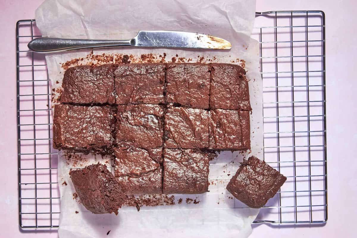 Nine chocolate brownies on parchment paper, with two pieces separated, sit on a cooling rack. A butter knife with chocolate crumbs rests beside the brownies on a light pink background.