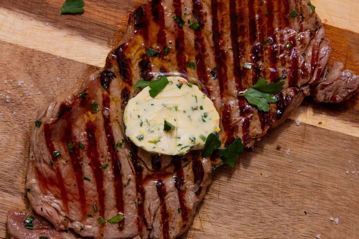 Grilled steak with visible sear marks topped with a round pat of herb butter and garnished with parsley, served on a wooden cutting board with a sprinkle of salt.