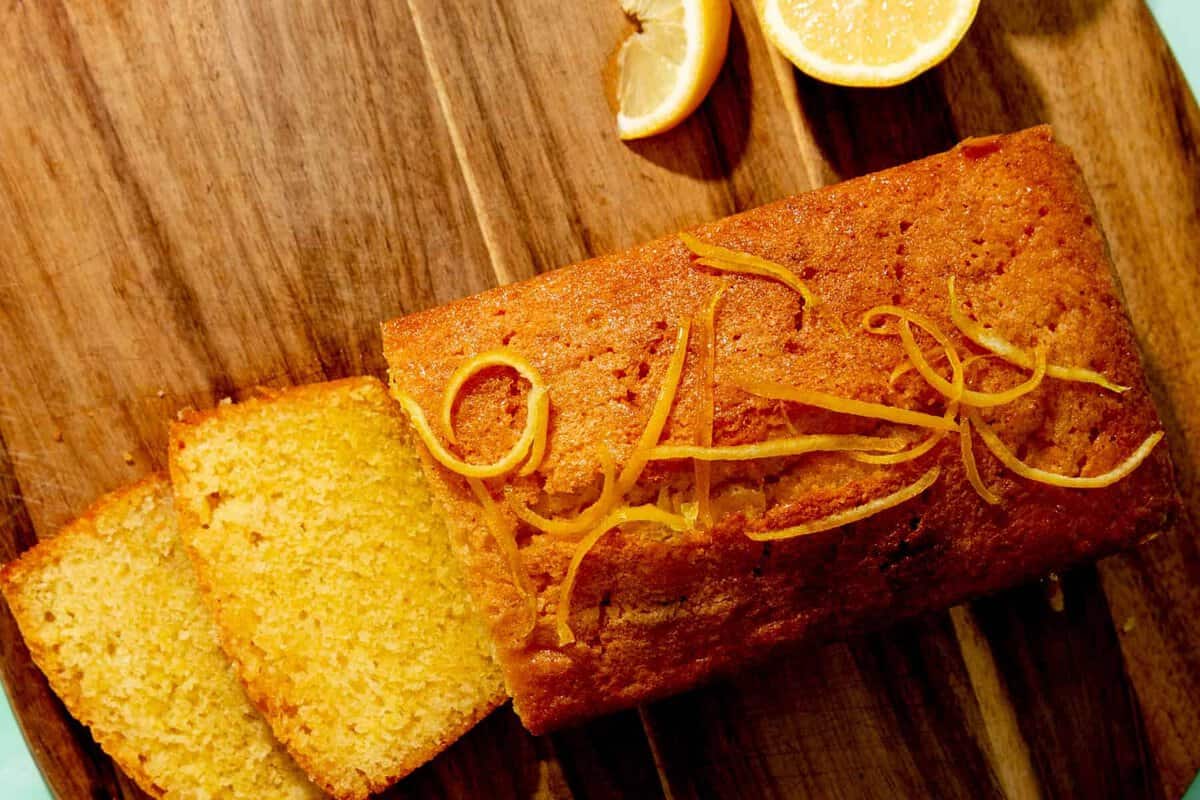 A loaf of lemon cake with lemon zest on top sits on a wooden board, with two slices cut and two lemon halves nearby.