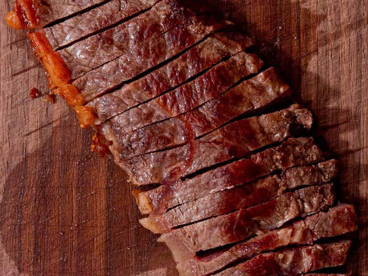 Sliced grilled steak arranged on a wooden cutting board, showing juicy, seared meat with a few pieces of fat along the edge.