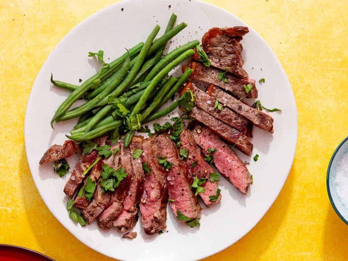 frozen steak in air fryer feature image of perfectly cooked steak that has been sliced and fanned around a white plate next to green beans and sprinkled with parsley