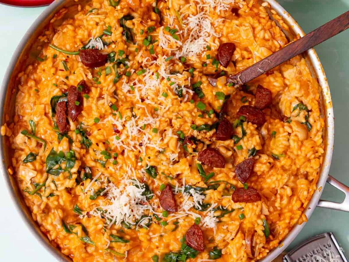 A large pan with choicken and chorizo risotto topped with parmesan, chorizo pieces and chives with a wooden spoon and small dishes of chives and chorizo next to the pan.