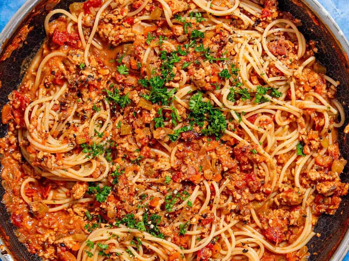 A skillet filled with cooked spaghetti mixed with a chunky tomato and ground meat sauce, garnished with chopped parsley, sits on a bright blue tiled surface.